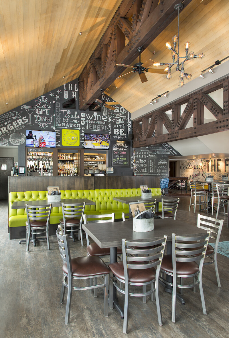 A modern restaurant interior with wooden beams, lime green booth seating, tables and chairs, and a counter area with multiple TV screens.