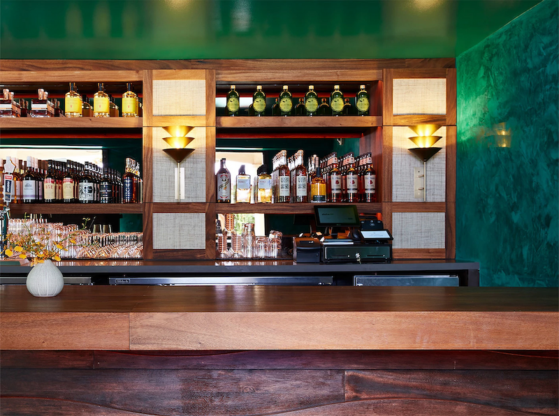 The image shows a well-organized bar with bottles of liquor on shelves, a cash register, and glassware; there's a small pumpkin on the countertop.