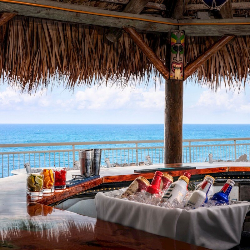 A tropical bar by the ocean with various bottles in ice, tiki decor, and a stunning sea view under a thatched roof structure.