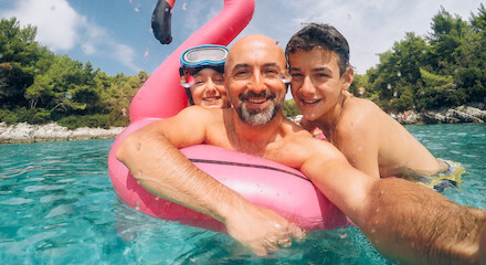 A man and two children are enjoying a swim, posing with a pink flamingo float in clear blue water, surrounded by trees.