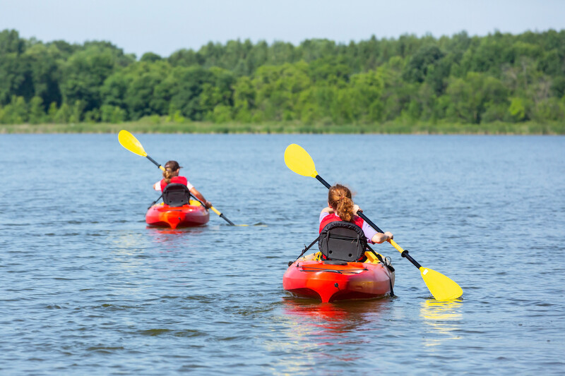 Two people are kayaking on a lake, each in a red kayak with yellow paddles, surrounded by lush green trees.