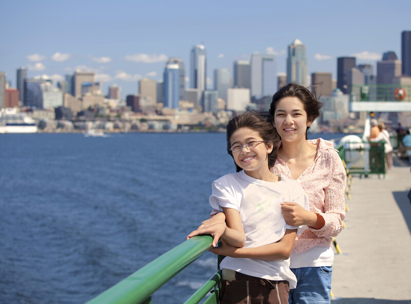 Two people, possibly siblings, smiling on a ferry with a city skyline in the background, likely Seattle due to the recognizable buildings.