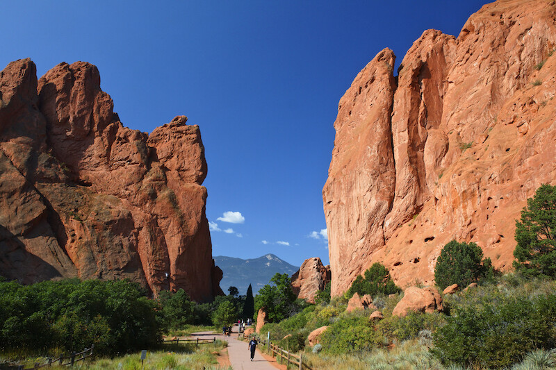 A scenic hiking trail flanked by tall red rock formations, with a clear blue sky and distant mountains in the background.