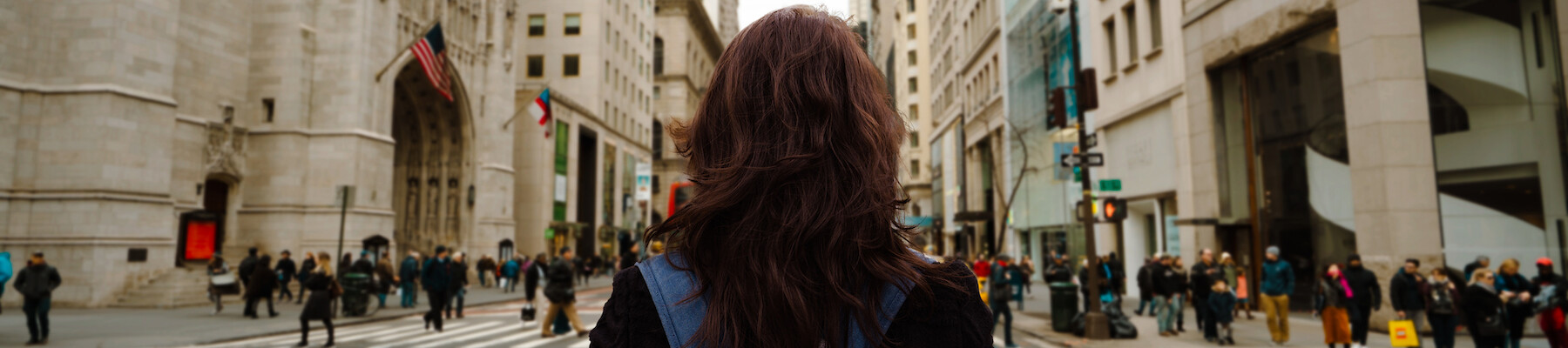 A person with a blue backpack stands in the middle of a busy city street, surrounded by tall buildings and pedestrians, facing away from the camera.