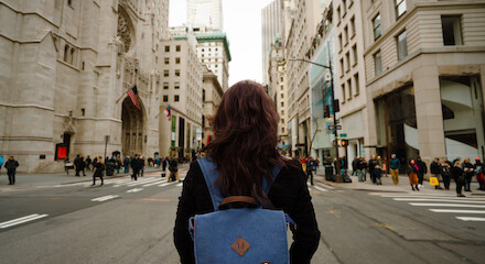 A person with a blue backpack stands in the middle of a busy city street, surrounded by tall buildings and pedestrians, facing away from the camera.