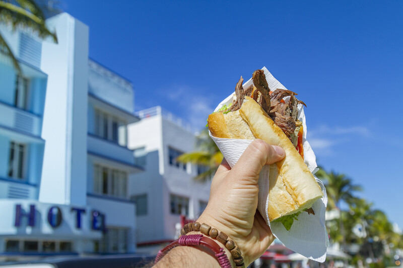 A hand holding a sandwich with meat and lettuce in front of a building labeled "HOTEL" under a clear blue sky with palm trees in the background.