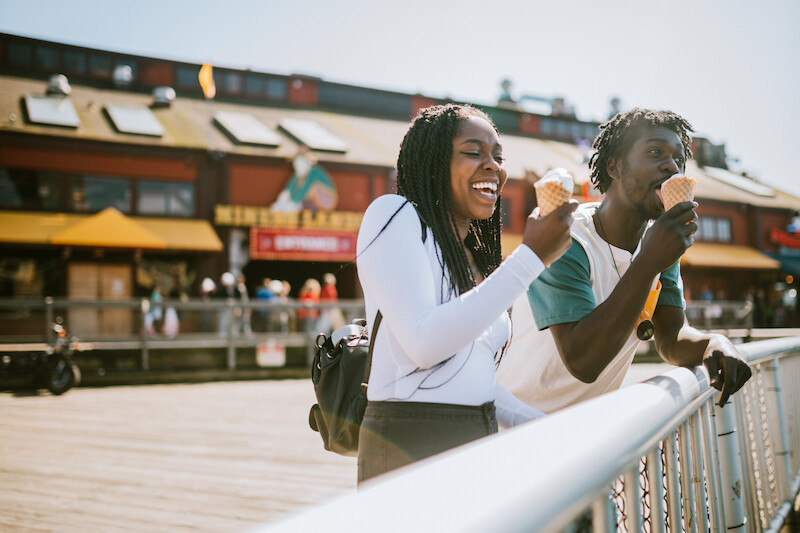 Two people are happily enjoying ice cream cones outside a building, standing by a railing, with a bright sunny day and a busy street in the background.