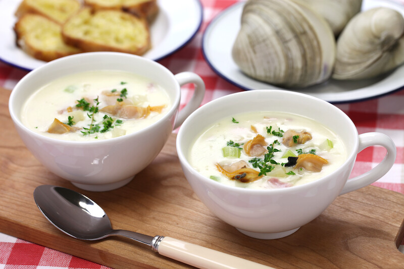 Two bowls of clam chowder garnished with herbs are on a wooden board with a spoon. Clams and bread slices are also present.