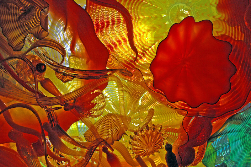 The image depicts a colorful arrangement of abstract, intricate glass sculptures in vivid hues of red, orange, yellow, and green.