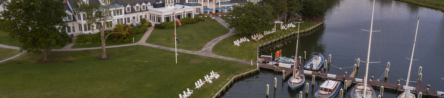 The image shows a scenic waterfront with boats docked at a marina. There's a large building surrounded by trees and a lawn with white chairs.