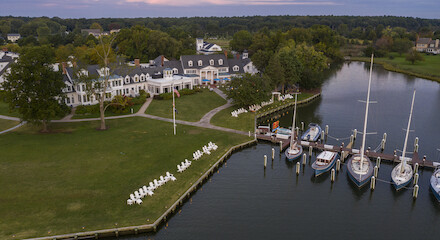 The image shows a scenic waterfront with boats docked at a marina. There's a large building surrounded by trees and a lawn with white chairs.