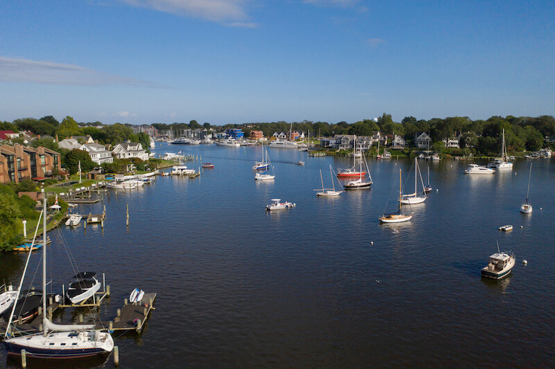 The image shows a scenic view of a calm harbor with various sailboats and small yachts anchored, surrounded by greenery and waterfront homes.