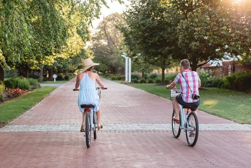 Two people are riding bicycles on a brick path in a park-like setting, surrounded by lush green trees and a neatly maintained landscape, ending the sentence.