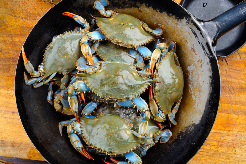 A pile of blue crabs arranged in a black skillet on a wooden surface, ready for cooking.