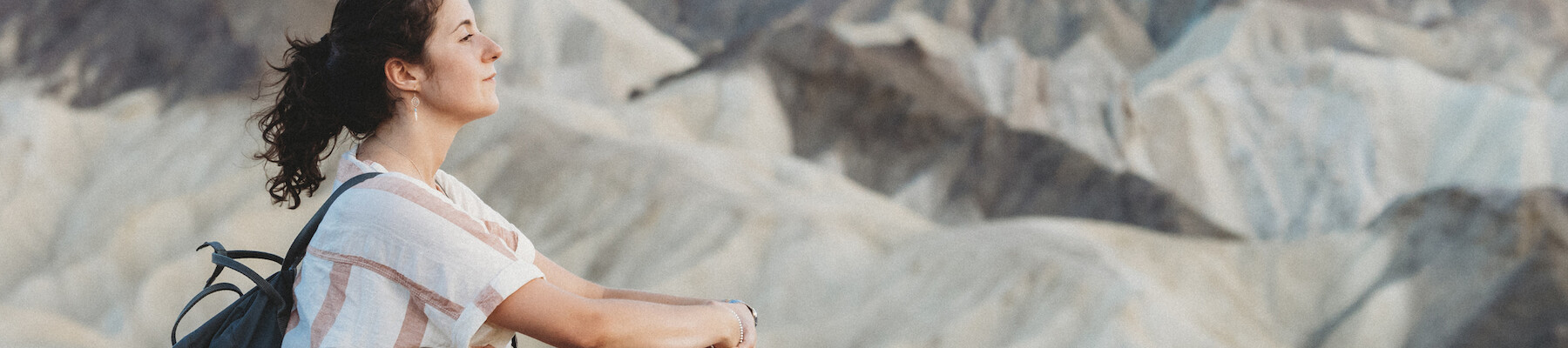 A person with a backpack sits on a rock ledge, facing a mountainous landscape, looking serene and contemplative.