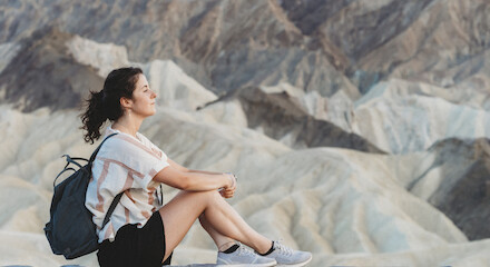 A person with a backpack sits on a rock ledge, facing a mountainous landscape, looking serene and contemplative.