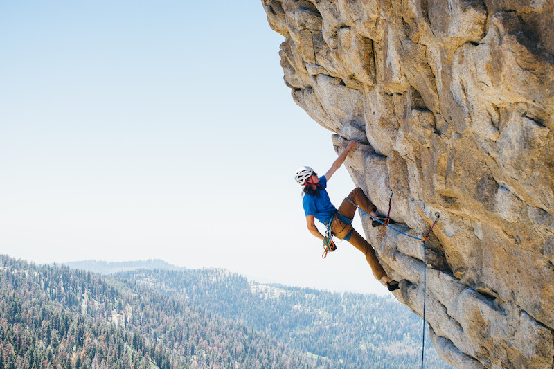 A climber wearing a helmet scales a steep rock face with mountains and forest in the background on a clear day, mid-climb.