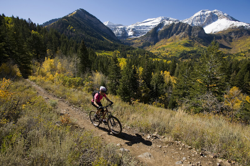 The image shows a person mountain biking on a dirt trail surrounded by forested hills and snow-capped mountains under a clear blue sky.