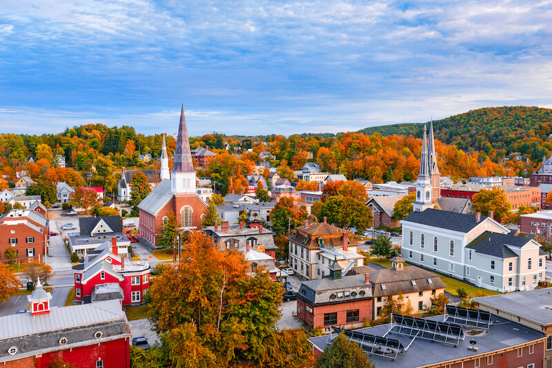 The image showcases a quaint town with colorful autumn trees, historic buildings, and churches with tall steeples, under a partly cloudy sky.