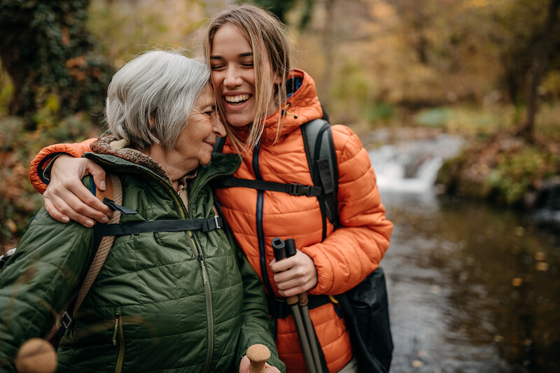 Two people in warm clothing, hiking and embracing by a river with a waterfall in the background. Both have hiking poles and are smiling happily.