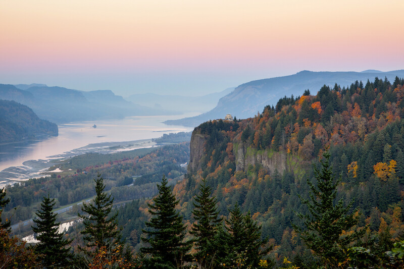 A scenic view of a river winding through forested mountains with a colorful sunset sky, featuring lush greenery and a cliffside.