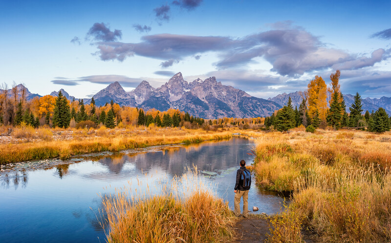 A person stands near a calm river surrounded by autumn foliage, looking at majestic mountains and partly cloudy skies in the background.