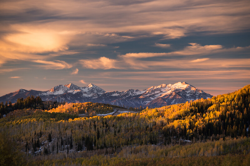 The image shows a picturesque landscape with snow-capped mountains under a partly cloudy sky and forested hills in autumn colors.