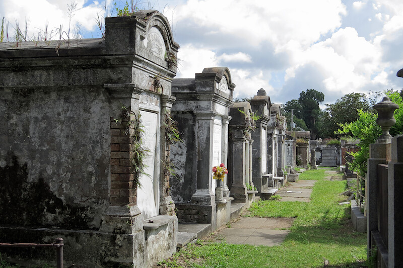 A row of weathered above-ground tombs in a cemetery, with a grassy path and some flowers, under a partly cloudy sky.