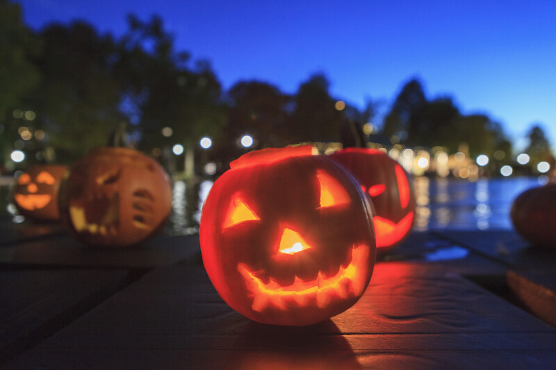 A glowing jack-o'-lantern with a carved smiling face sits on a table outdoors, with more pumpkins and blurred lights in the background.