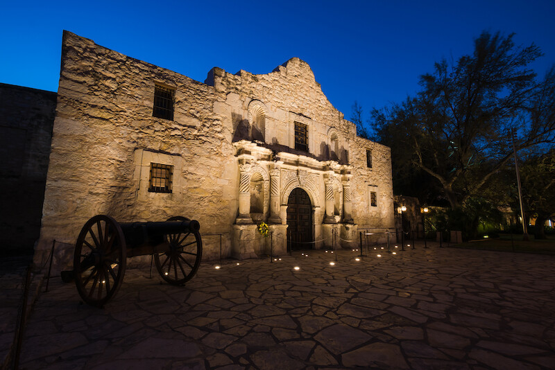 This is an image of a historic stone building with a cannon to the side, lit up against a twilight sky, surrounded by trees.