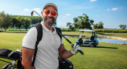 A man in golf attire is smiling, holding a golf club on a lush golf course. A golf cart and pond are visible in the background.