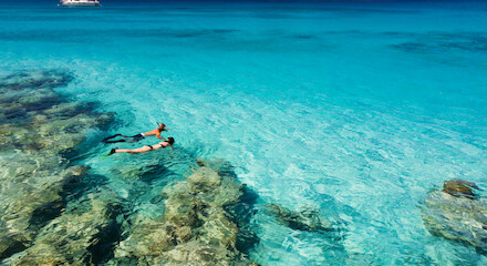 Two people snorkeling over a vibrant coral reef in clear turquoise water, with a sailboat and islands in the distant background.