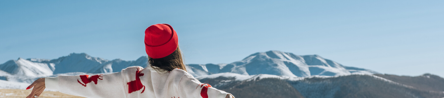 A person in a sweater with deer patterns and a red hat stands arms outstretched in a snowy landscape with mountains under a clear blue sky.