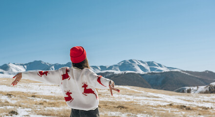 A person in a sweater with deer patterns and a red hat stands arms outstretched in a snowy landscape with mountains under a clear blue sky.