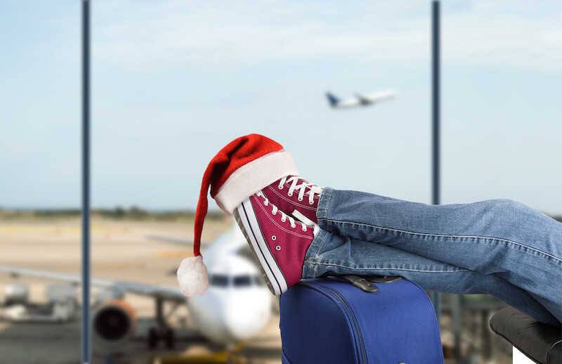 A person in jeans with red shoes and a Santa hat rests feet on a suitcase at an airport with planes outside.