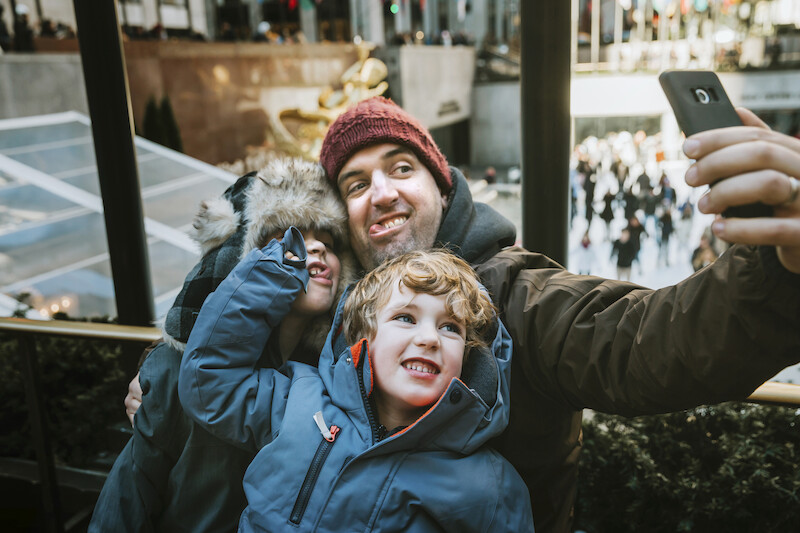 A person and two kids are bundled up in warm clothing, taking a selfie outdoors with a crowd and festive decorations in the background.