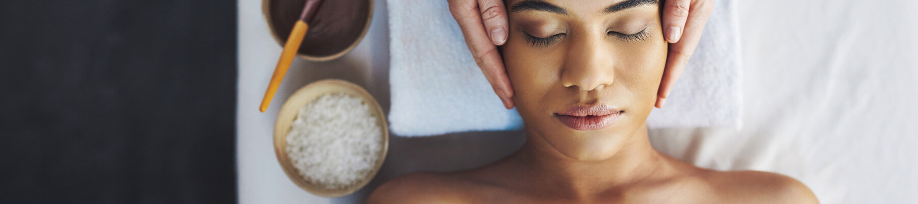 A person receives a facial massage, lying down with eyes closed. Nearby are bowls of oil, powder, and a brush on a white towel.