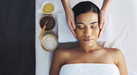 A person receives a facial massage, lying down with eyes closed. Nearby are bowls of oil, powder, and a brush on a white towel.