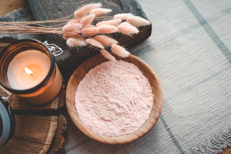 A candle, a bowl of pink powder, and dried flowers are arranged on a wooden surface, creating a cozy and serene atmosphere.