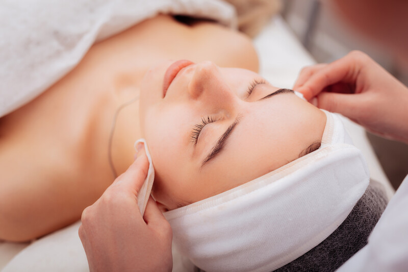 A person receiving a facial treatment, with a towel around their head, while someone adjusts cloths on their cheeks.