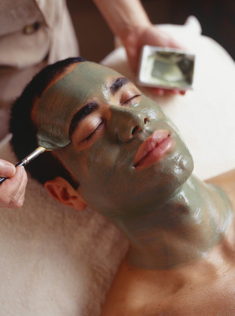 A person is lying down with a green facial mask being applied with a brush, enjoying a relaxing spa treatment.