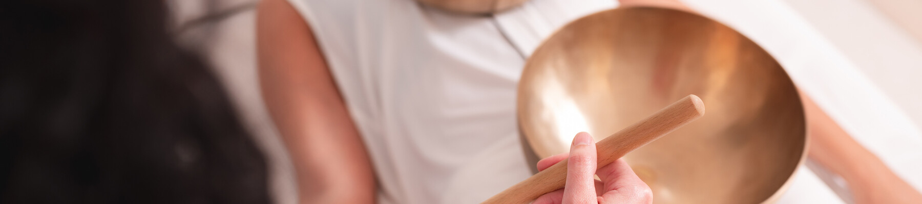 A person is lying down with singing bowls placed on them, while another person uses a mallet. This appears to be a sound healing session.