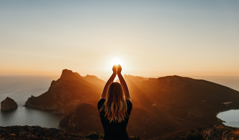 A person stands with arms raised towards the sun, overlooking a scenic coastal landscape during sunset.