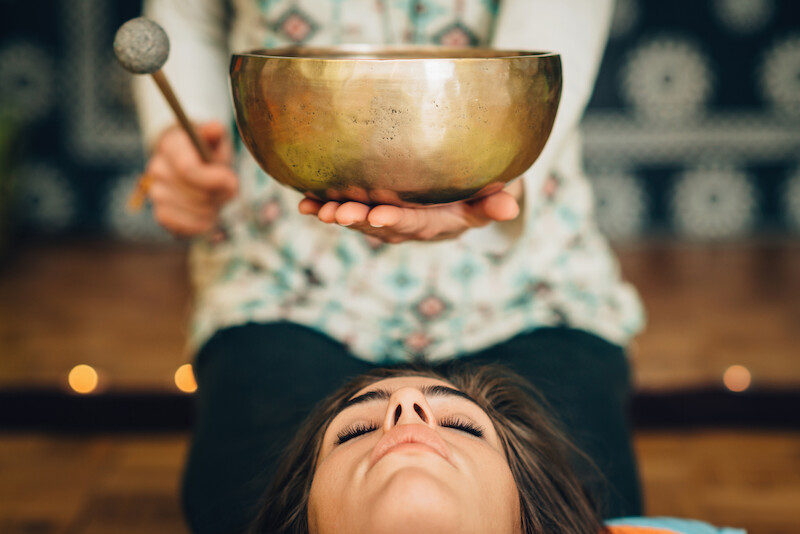 A person holds a singing bowl over a reclining individual's head, suggesting a sound therapy or meditation session.