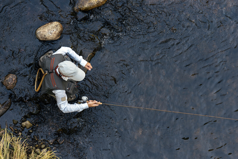 A person is fly fishing in a calm stream, surrounded by rocks and wearing a hat and vest.