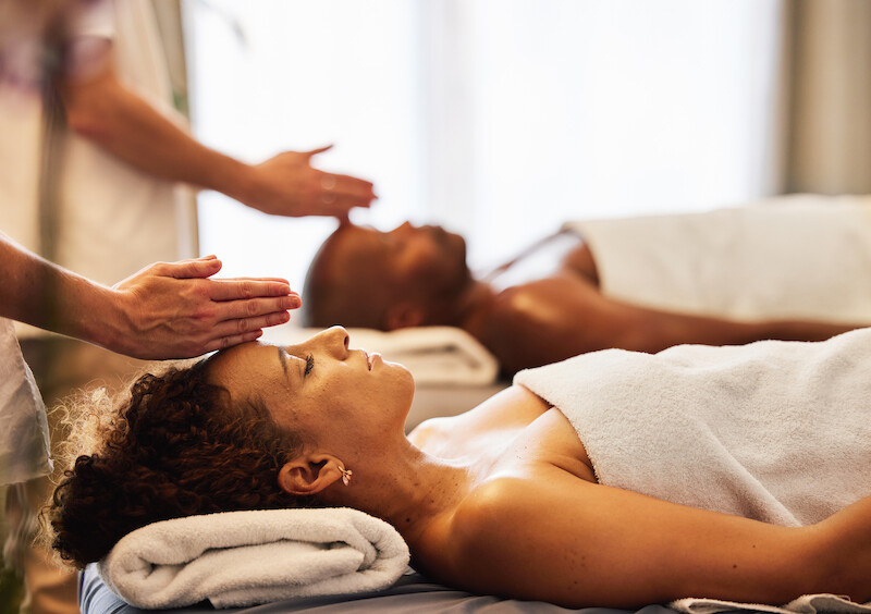 Two people are lying on massage tables, covered with towels, while receiving a forehead massage in a relaxing spa setting.