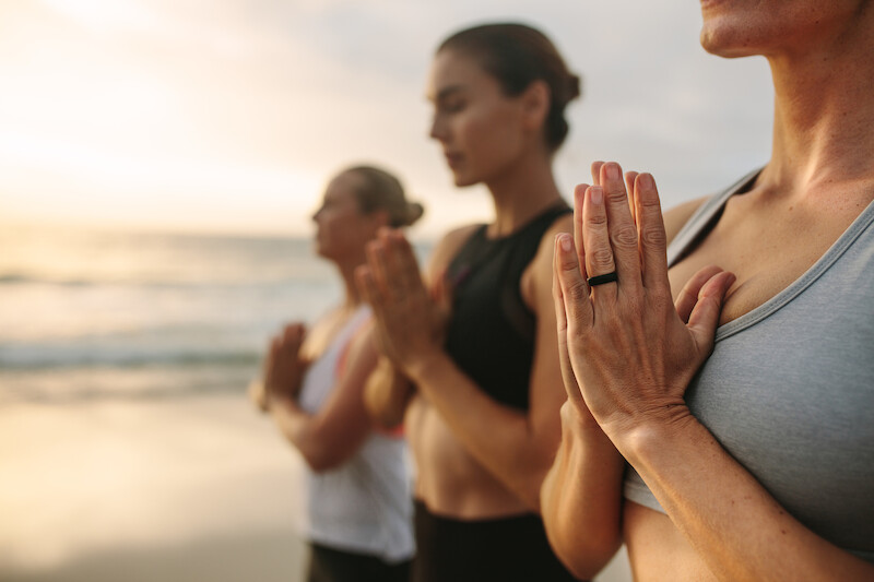 Group of people practicing yoga on a beach, standing with hands in prayer position, with the ocean in the background and a warm glow.