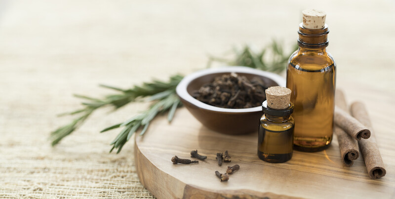 The image shows bottles of essential oil, cinnamon sticks, cloves, and herbs on a wooden surface.