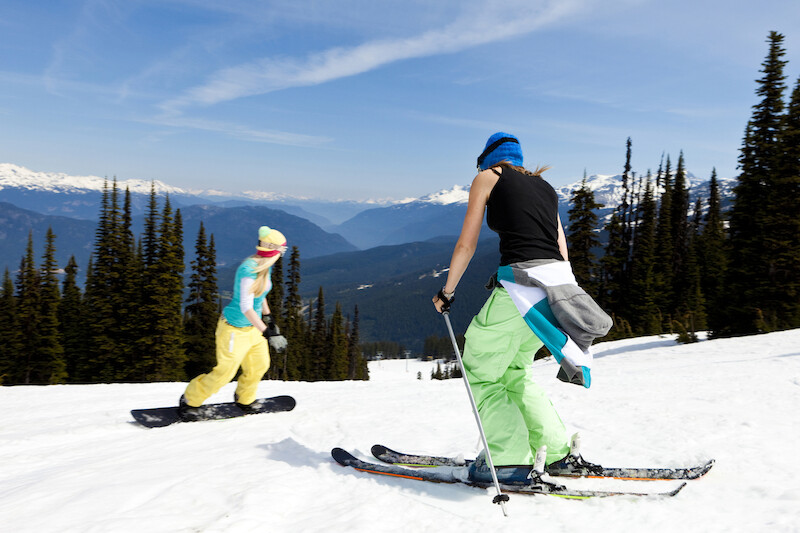 The image shows two people enjoying skiing and snowboarding on a sunny day in a snowy mountain landscape with trees.