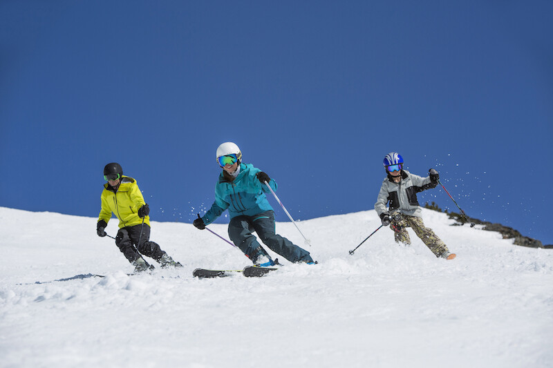 Three people are skiing on a snowy slope under a clear blue sky, with each wearing colorful jackets and ski gear.